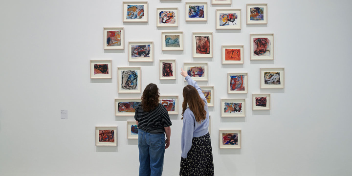Two individuals standing next to each other in a gallery space  at Kettle's Yard looking at artworks on a wall.