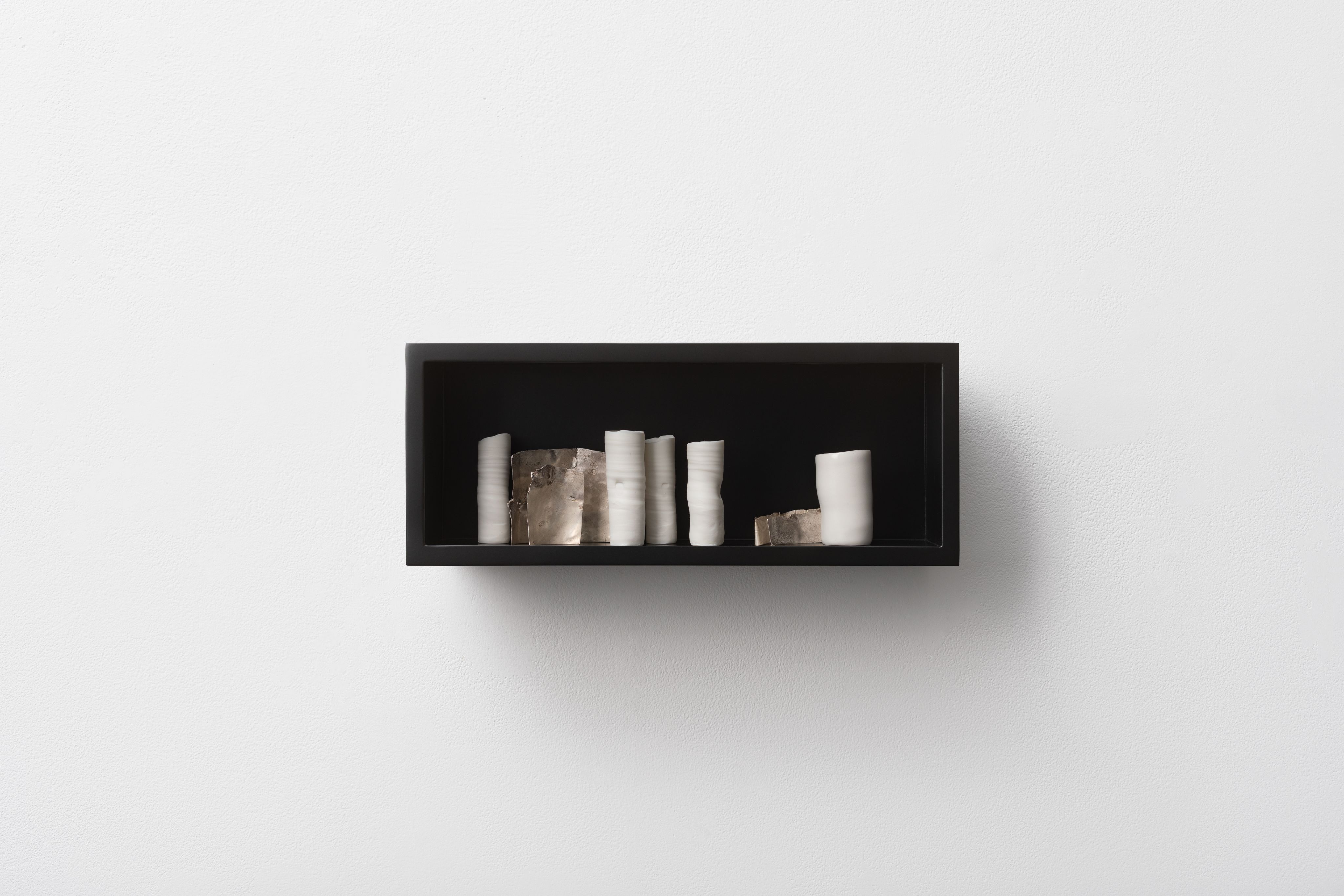 A black rectangular wall‑mounted shelf displaying a row of small ceramic forms in pale and neutral tones, set against a white textured wall.