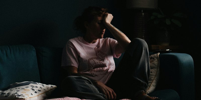 Woman sitting on sofa in the dark, placing a hand to her forehead.